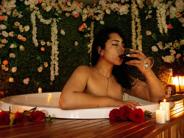 ScarlettBigAss, sem blusa, em uma banheira de hidromassagem, tomando vinho tinto, com rosas vermelhas e velas, segurando um copo, cabelo cacheado escuro, pele bronzeada, seios pequenos, relaxada, sensual..