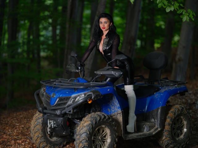 KarinaGray met zwarte laarzen, een zwarte latex outfit en een roze top, zittend op een blauwe, modderige quad, haar billen omhoog, handen op het stuur, met een bos op de achtergrond..