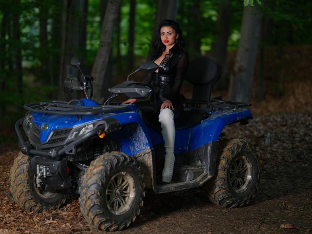 KarinaGray, vêtue d'une tenue en cuir noir et chaussée de bottes blanches, assise sur un VTT bleu sale, avec un arrière-plan de forêt, cheveux noirs, rouge à lèvres rouge, regardant la caméra. Les seins sont visibles sous la tenue..