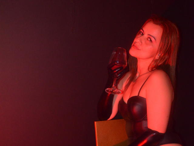 Perla Divaio, con cabello rojo, vestido de cuero negro y guantes, sosteniendo una copa de vino tinto, sonriendo levemente, sentada en una iluminación roja tenue..