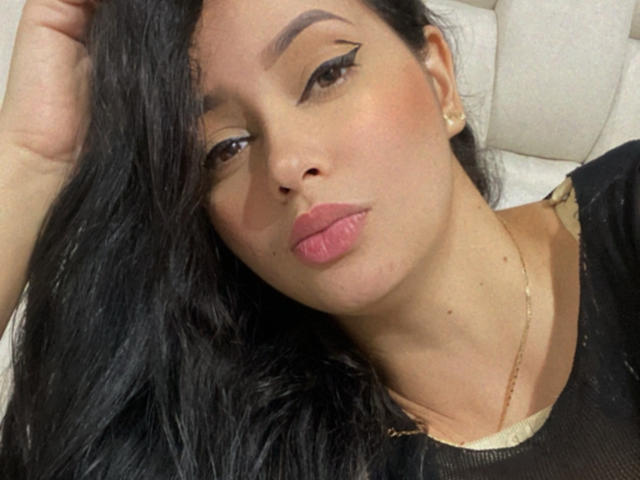 EmilyParkerr has long black hair, brown eyes with winged eyeliner, pink lips, and a gold necklace. She's looking at the camera with a slightly pouty expression.