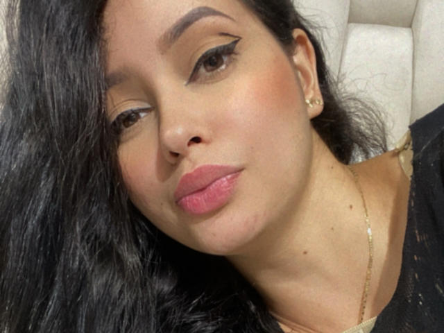 EmilyParkerr's face with long black hair, pink lips, and gold eyeliner. She's wearing a gold necklace and black top, with a small mole on her left cheek.