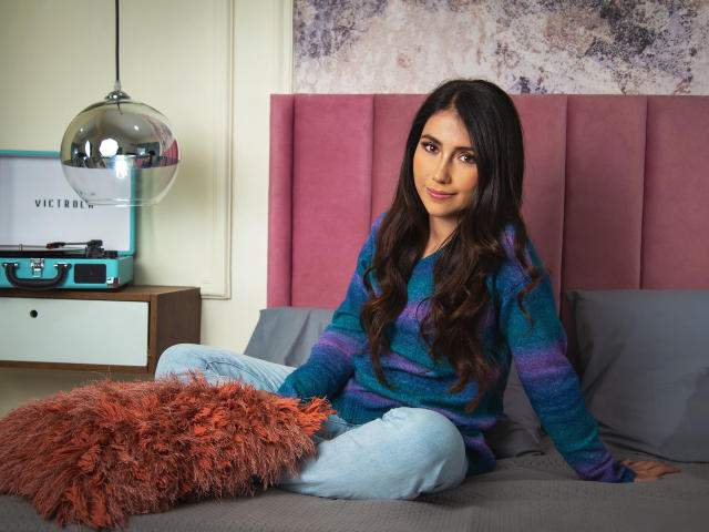 Alana Bazan, avec de longs cheveux foncés, portant un pull rayé bleu-violet et un jean bleu clair, assise sur un lit avec un oreiller orange moelleux, regardant l'appareil photo..