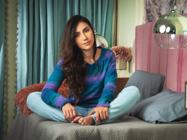 AlanaBazan with long brown hair, wearing a blue-purple striped sweater and light blue pants, sitting cross-legged on a bed, looking at the camera, barefoot, with a calm expression.