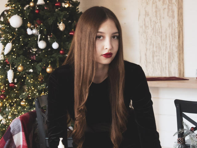 EllyCat con lunghi capelli castani, rossetto rosso, top di velluto nero, in posizione leggermente inclinata in avanti, albero di Natale con decorazioni sullo sfondo, seduta su una sedia nera, espressione seria, carina, sexy..