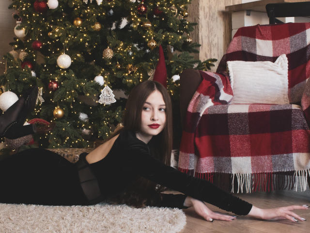 EllyCat, con piel pálida y cabello largo castaño, está acostada sobre una alfombra esponjosa, vestida con un vestido negro, estirándose, con un árbol de Navidad y una manta de cuadros rojos en el fondo..