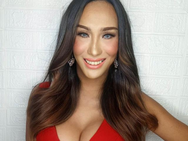 WOWPinayWilFrid, com cabelos castanhos longos e ondulados, olhos azuis, vestido vermelho e decote, sorrindo para a câmera, usando brincos elaborados, tem uma aparência fofa e sexy..