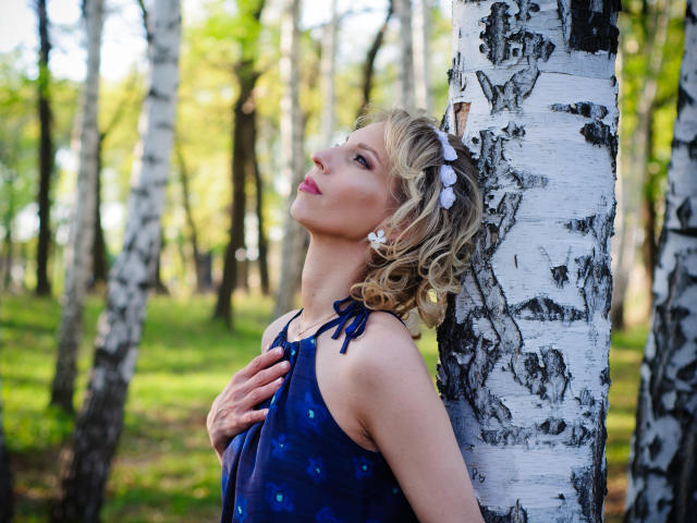 AdrianaSea leans against a birch tree, wearing a blue dress with blue flower patterns, blonde curly hair with a white flower, hand on chest, looking up, tits visible.