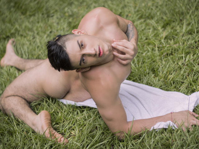 Jonathan Jeremiah, nackt, liegt auf dem Gras mit einem weißen Handtuch, den Kopf geneigt, ein Arm bedeckt die kleinen Brüste, kurze schwarze Haare, ein Tattoo am Arm, blickt in die Kamera, rote Lippen, nackte Brust..