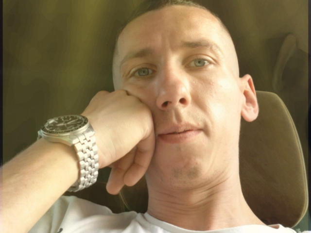 Trojanos199328, met kort bruin haar, blauwe ogen en een licht geopende mond, leunt met zijn kin op zijn hand, draagt een zilveren horloge en een wit shirt..