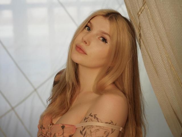 Aleksa Keller, con lunghi capelli biondi, pelle chiara e piccoli seni, indossa una maglietta rosa con fiori, guarda verso l'alto con un'espressione dolce, ed è in piedi vicino a una tenda..