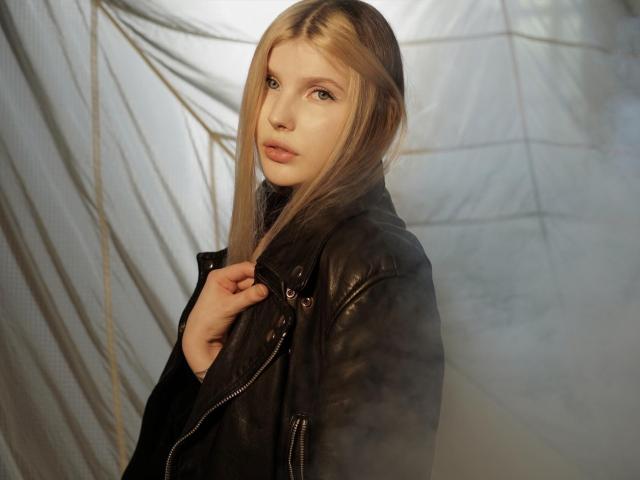 AleksaKeller with long blonde hair, wearing a black leather jacket, looking over shoulder with a slight smile, holding jacket collar, soft light, subtle makeup, gentle expression.