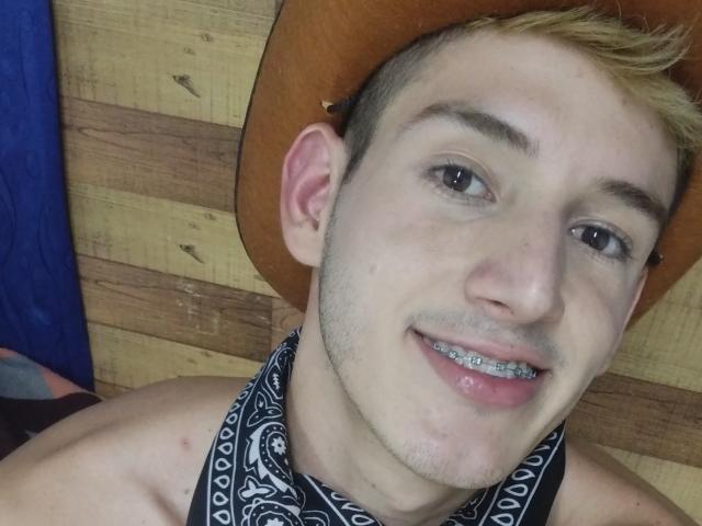 JonathanJeremiah has short blonde hair, a brown hat, and a black and white bandana, with silver metal teeth and a slight smile, showing a cute, nerdy face.