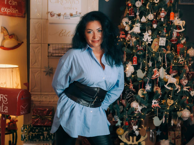 Anelisa Reys mit welligem, schwarzem Haar, einem blauen Hemd, einem schwarzen Gürtel und einer schwarzen Hose, steht neben einem geschmückten Weihnachtsbaum, lächelt und zeigt ihr Gesäß, wobei ihre Hände hinter ihrem Rücken sind..