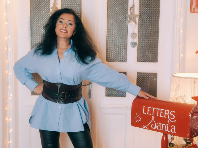 Anelisa Reys, con cabello oscuro y rizado, sonriendo, vestida con una camisa azul abotonada, un cinturón negro y pantalones de cuero negro, apoyada en un buzón rojo con la etiqueta "Cartas a Papá Noel".."