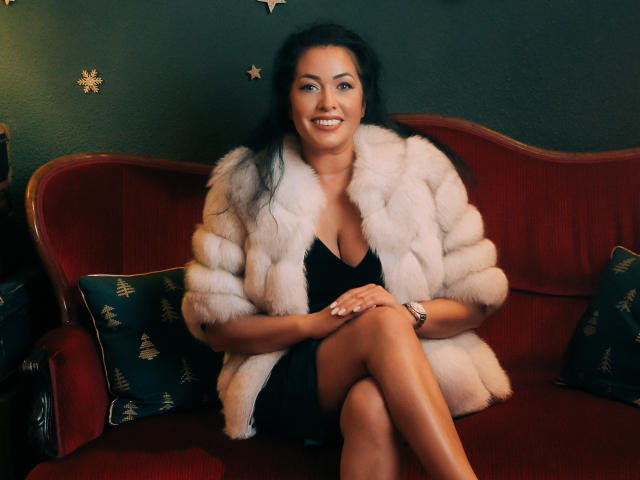 Anelisa Reys, mit gebräunter Haut, schwarzen Haaren und einem Lächeln, trägt einen flauschigen weißen Pelzmantel, ein schwarzes Kleid und eine goldene Uhr und sitzt auf einem roten Sofa mit grünen Kissen..