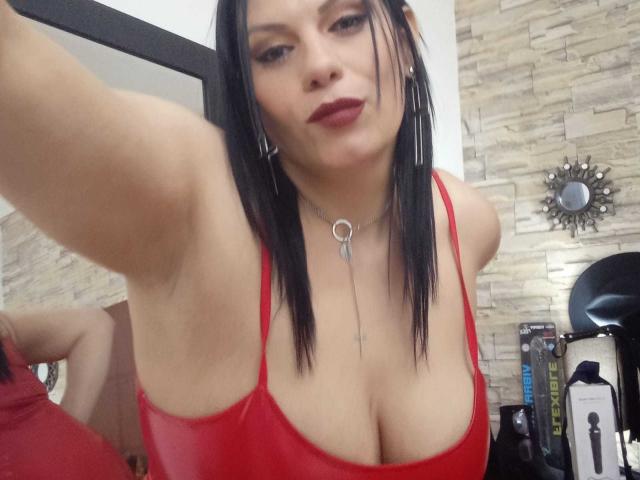 LizaRussell with black hair, red lipstick, and red top showing big tits, wearing a key necklace, taking a selfie in a mirror. Her arm is raised, eyes slightly down.