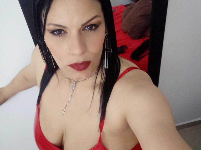 LizaRussell with black hair, red lipstick, and red bra, wearing a cross necklace, looking up with sultry eyes, showing her big tits, in a mirror selfie.
