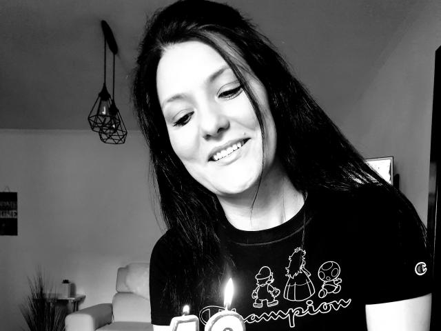 JoliDanielle with long dark hair, smiling at a candle she's holding. She's wearing a black shirt with white line drawings. Black and white photo, looking cute.