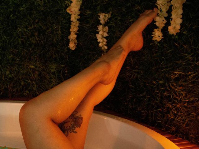 As pernas de ScarlettBigAss estão em uma banheira branca, sua pele brilhante, uma tatuagem na coxa direita, os pés apontados para cima, guirlandas de flores brancas penduradas acima..