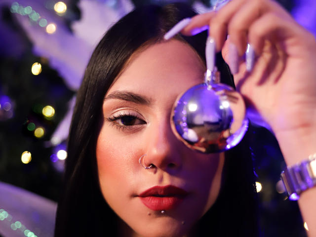 HaleyBeckham with dark hair, nose ring, and lip piercing, holding a shiny silver ball in front of her face, wearing red lipstick, and white nail polish.