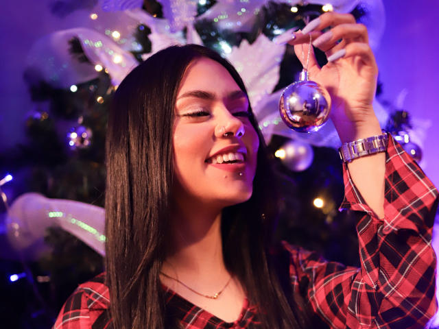 Haley Beckham, avec de longs cheveux noirs, souriante, tenant un ornement de Noël argenté brillant, portant une chemise à carreaux rouges, un anneau au nez et un collier en or, dans un décor illuminé de violet..