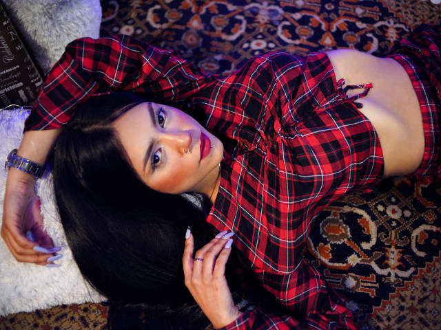 HaleyBeckham with black hair, red plaid shirt, and matching pants, lying on a rug, showing her tits, wearing a bracelet, looking at the camera with red lipstick.