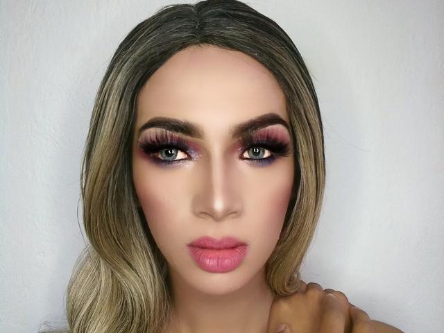 Het gezicht van CristalKlein, met blond haar, roze lippen en dramatische, donkere oogmake-up. Haar hand rust op haar schouder. Ze heeft blauwe ogen en volle wenkbrauwen..