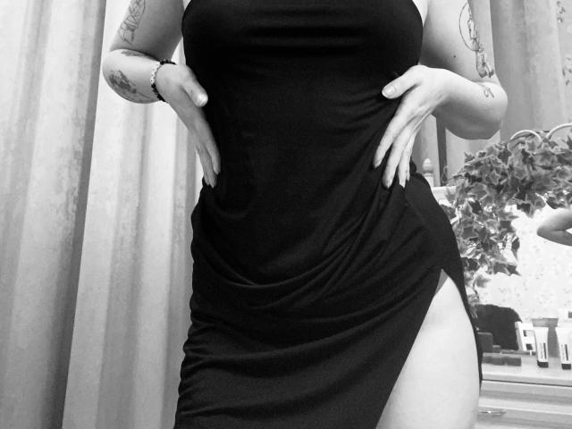 Black dress, KarKutie's hands on hips, showing a thigh slit, tattoos on arms, no face visible, black and white, KarKutie's tits slightly visible, standing, hands on dress.