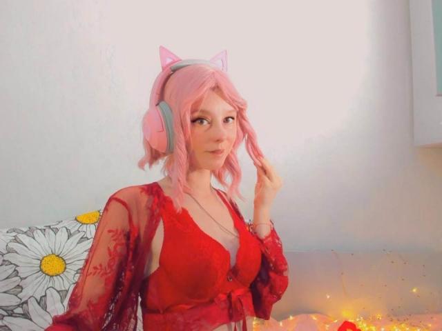 MissAstravert com orelhas de gato cor-de-rosa, cabelo ondulado cor-de-rosa, blusa de renda vermelha, mostrando seus pequenos seios, usando fones de ouvido cor-de-rosa, sentada com luzes de fada brilhantes e um travesseiro com padrão de margaridas..