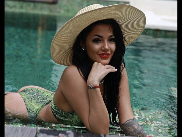 Yuukenzi, usando um biquíni verde com estampa de cobra, deitada em uma piscina, com cabelo preto molhado, batom vermelho e um chapéu de palha grande, mostrando o bumbum e o braço tatuado..