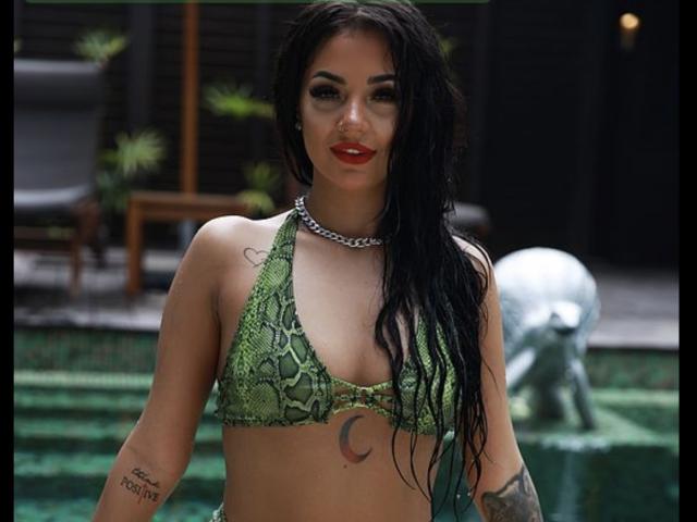 Yuukenzi with wet black hair, green snake-print bikini, red lipstick, chain necklace, tattoos, and a crescent moon tattoo on her chest, standing by a green-tiled pool.