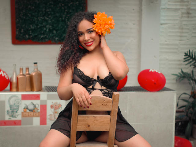 SelynRobert, con cabello rizado y una flor naranja en el pelo, lleva lencería de encaje negro, está sentada en una silla de madera, sonriendo, mostrando sus grandes senos, con lápiz labial rojo, con una mano en la cabeza, y hay botellas marrones en el fondo..
