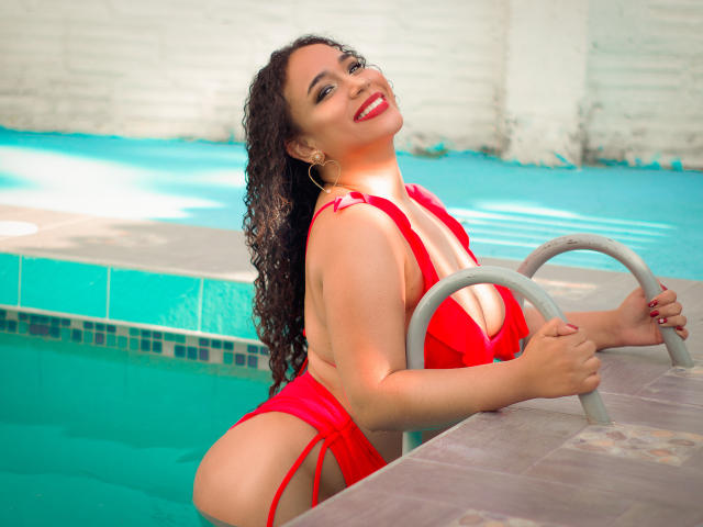 SelynRobert, con cabello oscuro y rizado, un bikini rojo y lápiz labial rojo, sonriendo junto a la piscina, sosteniendo la escalera, mostrando su gran trasero y sus pequeños pechos, y llevando aros de aro..