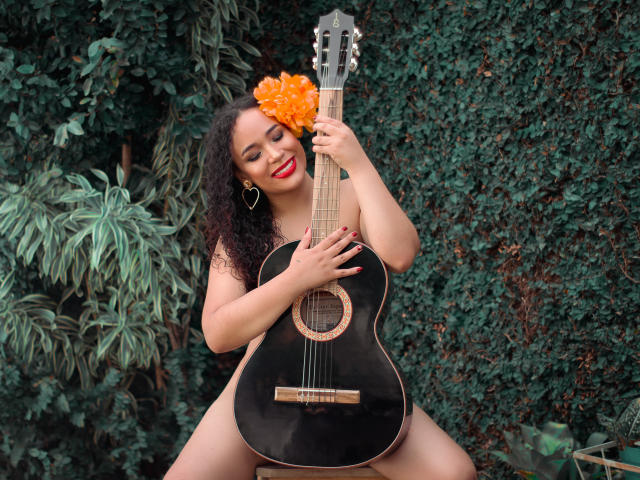 SelynRobert, con cabello oscuro y rizado, lápiz labial rojo y aretes con forma de corazón, sosteniendo una guitarra negra, con una flor naranja brillante en el cabello, sonriendo, sentada con parte de su cuerpo visible..