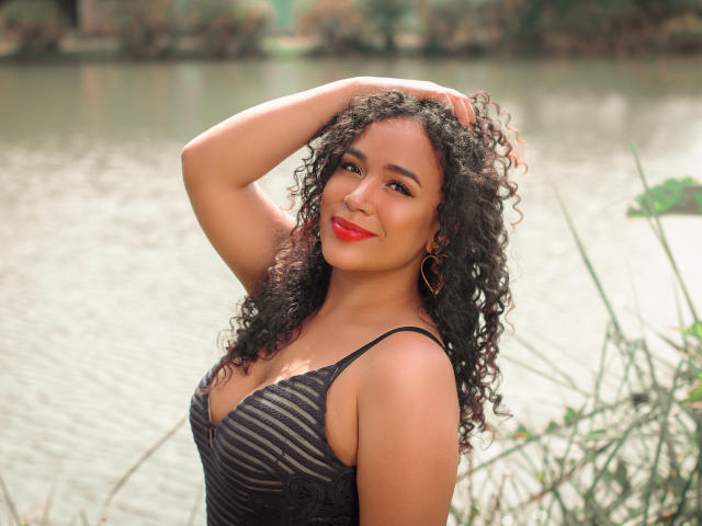 SelynRobert, con cabello negro rizado, lápiz labial rojo y aretes dorados, vestida con un vestido a rayas negras, con la mano en el cabello, de pie junto a un lago, sonriendo y mostrando sus senos..