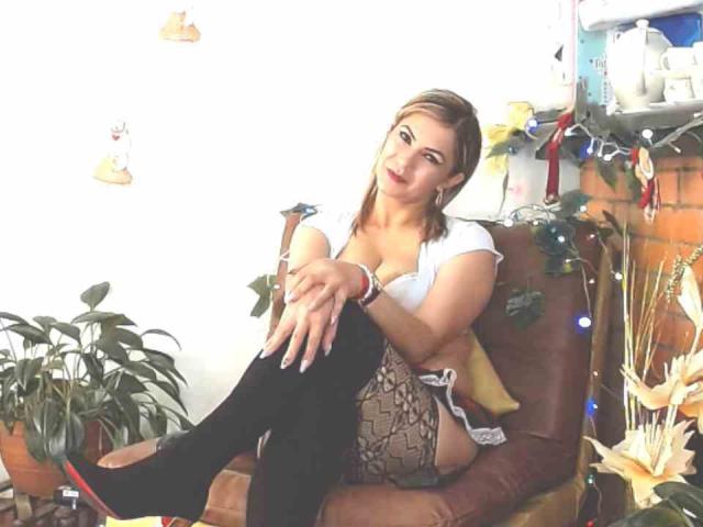 ChantalHotX, con senos pequeños, sentada en una silla marrón, vestida con una blusa blanca, medias negras hasta el muslo y una falda estampada, mirando a la cámara con lápiz labial rojo..
