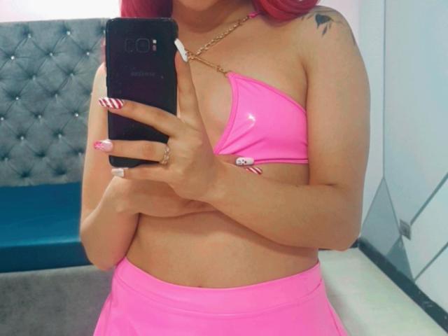 ValeryHarllow in a pink bikini top with small boobs, pink skirt, holding a phone, red nails, tattoo on right shoulder, gold necklace, in a mirror selfie.
