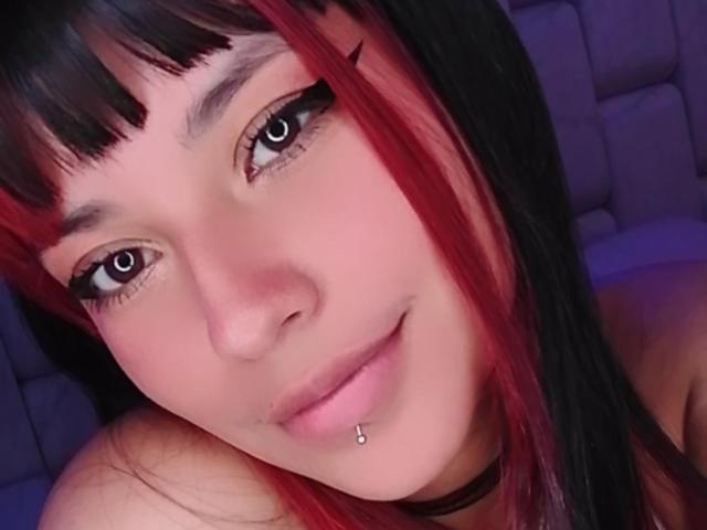NicolSanchez, avec des cheveux noirs et rouges, une frange droite, du crayon noir pour les yeux, des yeux foncés, une peau claire, un piercing au nez, un piercing aux lèvres, un léger sourire, portant un collier noir, allongée..