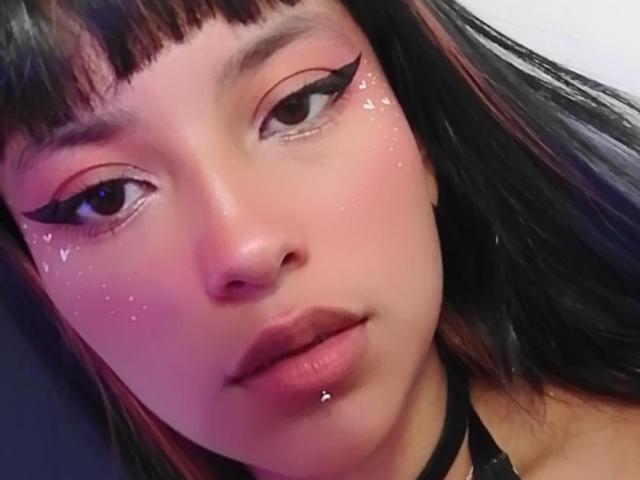 El rostro de NicolSanchez con maquillaje rosa en los ojos, delineador negro y purpurina brillante en las mejillas. Tiene el cabello negro con reflejos rosas, un piercing en el labio y un collar negro..