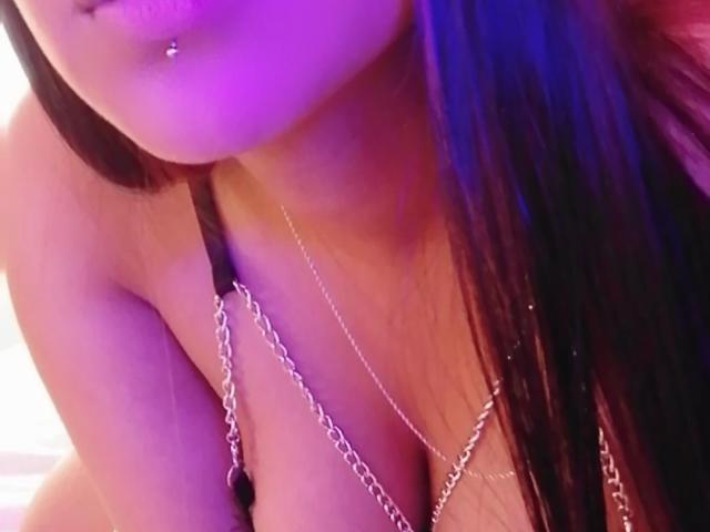El rostro de NicolSanchez, con un piercing en el labio, vistiendo un sostén negro y una cadena de plata cruzada sobre sus pechos, con iluminación púrpura y azul. Su cabello negro y lacio cubre parcialmente su pecho..