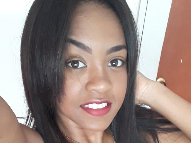 TiffanySammy has dark brown eyes, thick eyebrows, and red lipstick, with long black hair, a gold necklace, and a slightly open mouth showing white teeth, holding her hair up.