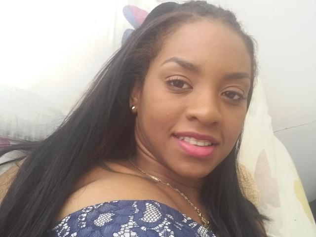 TiffanySammy has dark brown skin, long black hair, pink lipstick, small pearl earrings, and wears a blue patterned off-shoulder top. Her nipples are not visible.