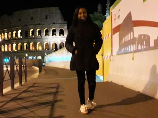 TiffanySammy with dark hair, black coat, black pants, and white sneakers, standing on a night-time sidewalk, hands on hips, smiling.