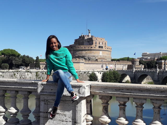 TiffanySammy, with black hair and green sweater, sits on a white balustrade, legs crossed, showing her ass, in front of a historic, circular stone building, clear blue sky.