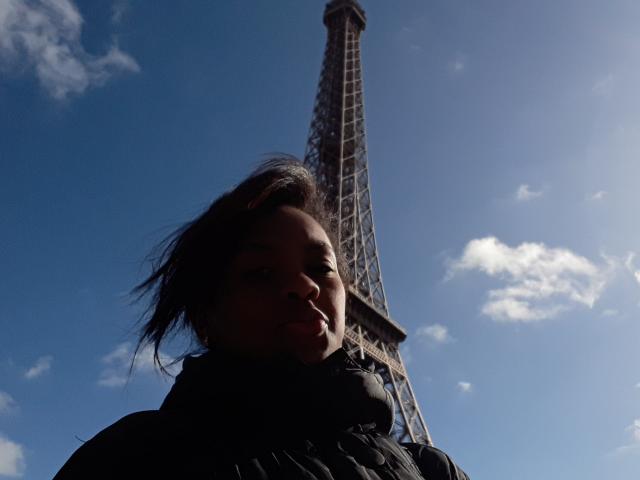 TiffanySammy with dark, windswept hair, black puffy jacket, and shadowed face, standing in front of the Eiffel Tower under a bright blue sky with small clouds.