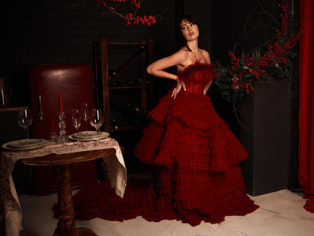 NinaTender in a red ruffled dress, hands on hips, standing by a table with red candles and wine glasses, black hair, red flowers, dark brick wall background.