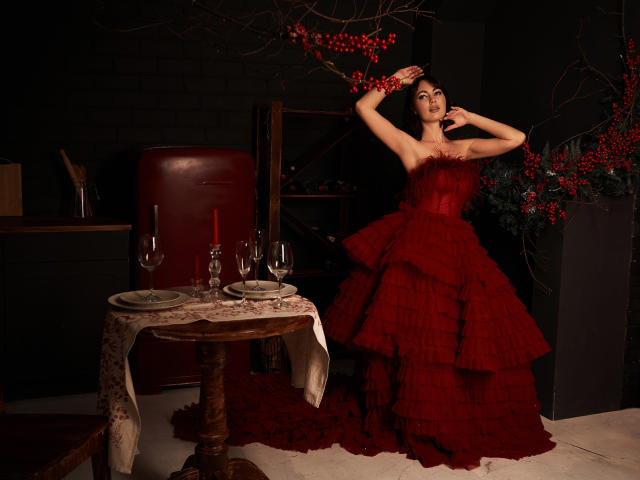 NinaTender vestindo um vestido vermelho, com babados, sem alças e com várias camadas, posando com os braços levantados, ao lado de uma mesa com copos de vinho e uma vela vermelha. Seus seios estão visíveis..