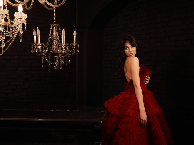 NinaTender in a red, ruffled, strapless dress with feathery trim, looking over her shoulder, standing beside a black, shiny, piano, under a crystal chandelier. Boobs visible.