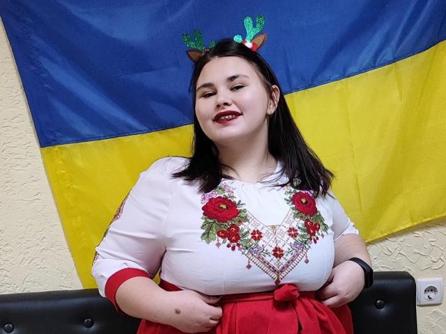 KaidoKun, mollig mit großen Brüsten, trägt ein weißes Oberteil mit roter Blumenstickerei, einen roten Rock und einen grünen Stirnband mit Geweihen, lächelt, hat dunkles Haar, eine weiße Wand im Hintergrund, eine blau-gelbe Flagge..